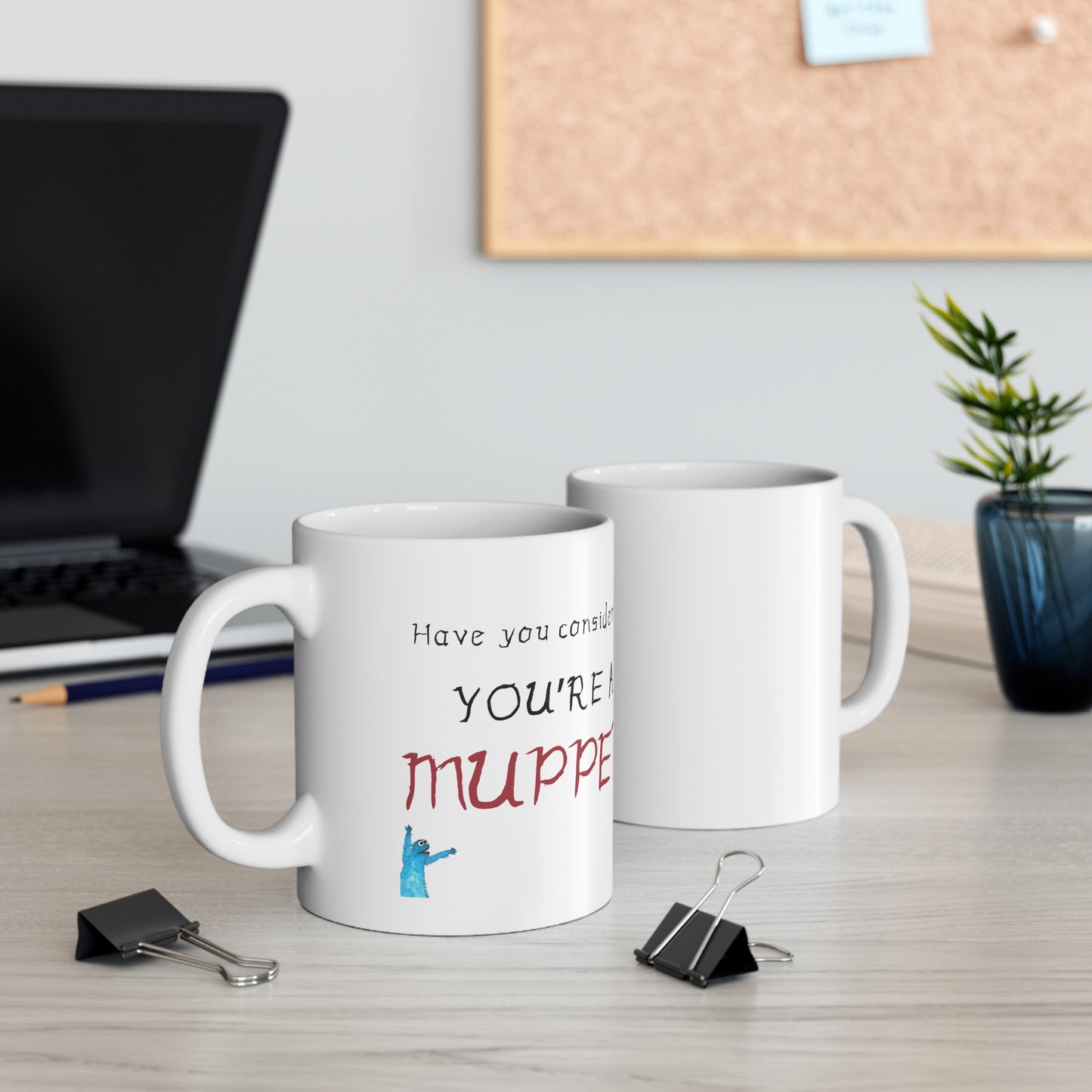 Two cups of white Coffee Mug with "Have you considered? You're a Muppet!" text and  muppet icon on a desk with laptop in back.